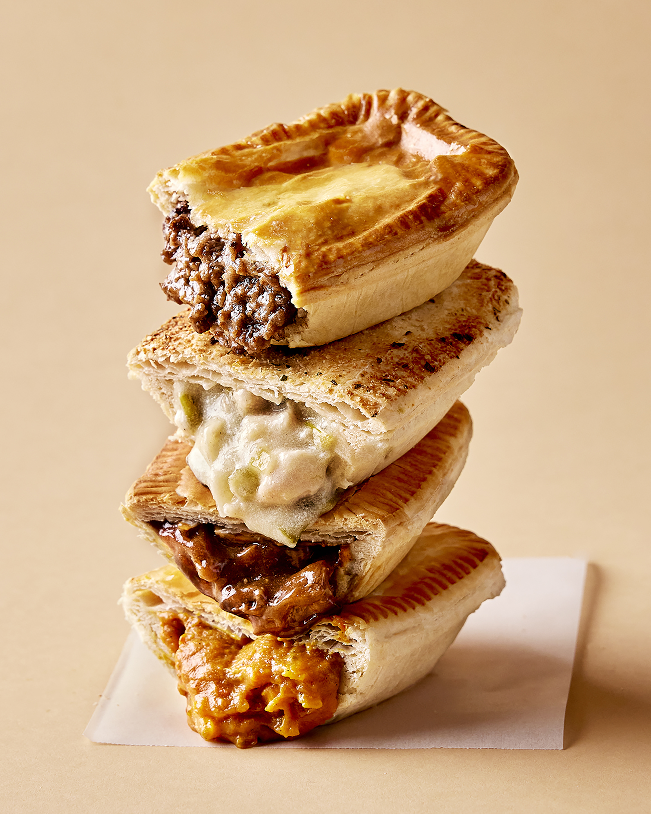 Tower of four savory meat pies in a vertical composition against a light brown backdrop, showcasing a variety of fillings and golden crusts.