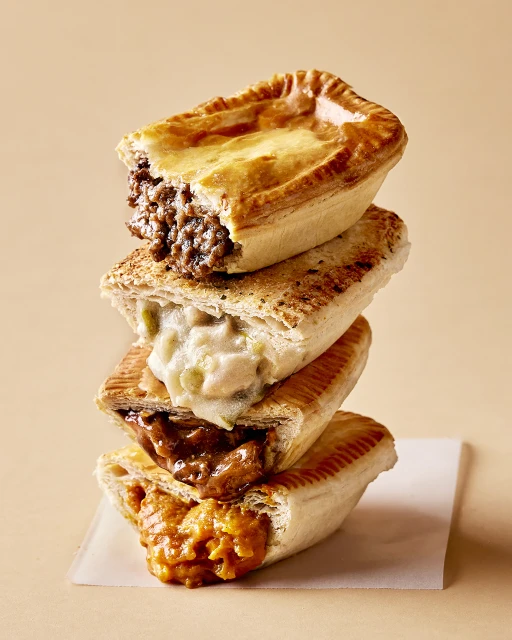 Tower of four savory meat pies in a vertical composition against a light brown backdrop, showcasing a variety of fillings and golden crusts.