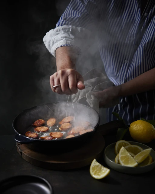 A person prepares food in a cast iron skillet, cooking sausage slices with steam rising, while a plate of lemon wedges sits nearby.