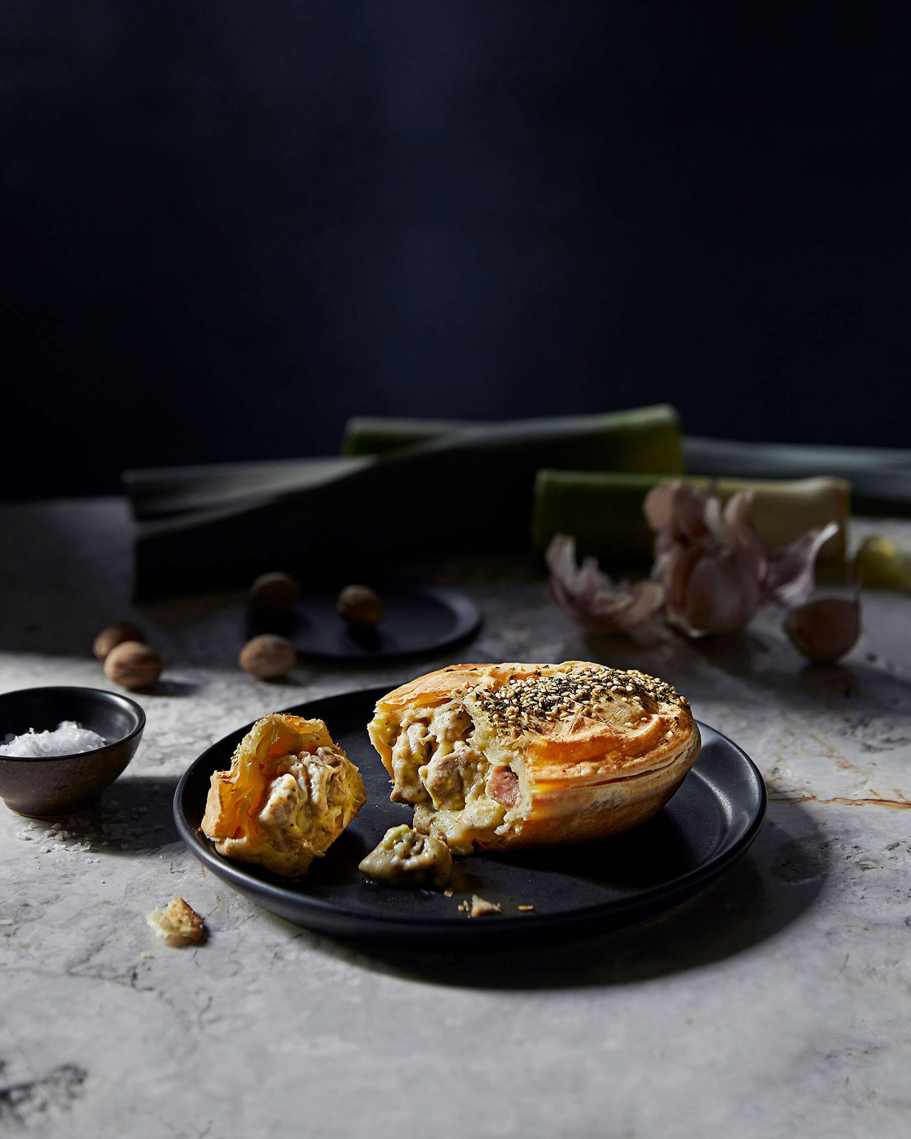 Studio shot of a savory meat pie on a black plate, partially cut open, with garlic cloves and nutmeg in the background, food photography.