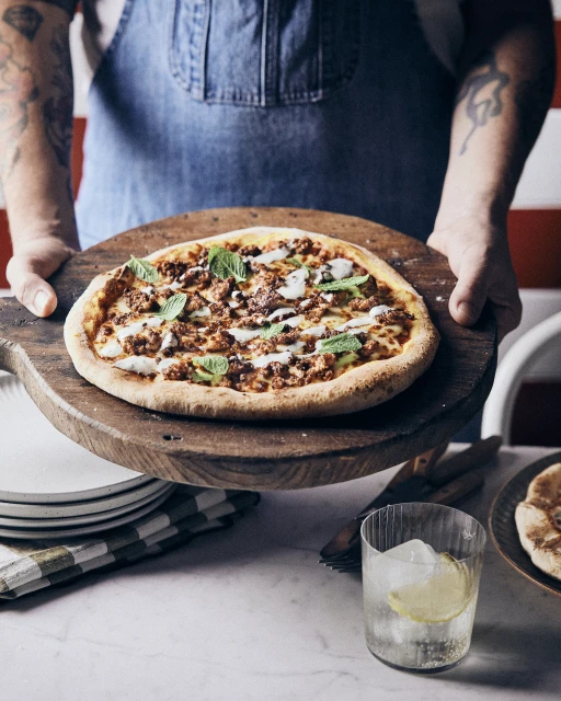 Overhead shot of a person holding a wood pizza peel with a pizza with green herbs, plates, and a drink on a white table.