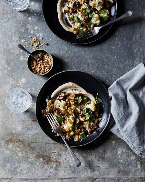 Overhead shot of two plates of roasted cauliflower salad with chickpeas, herbs, and a tahini dressing on a textured gray surface.