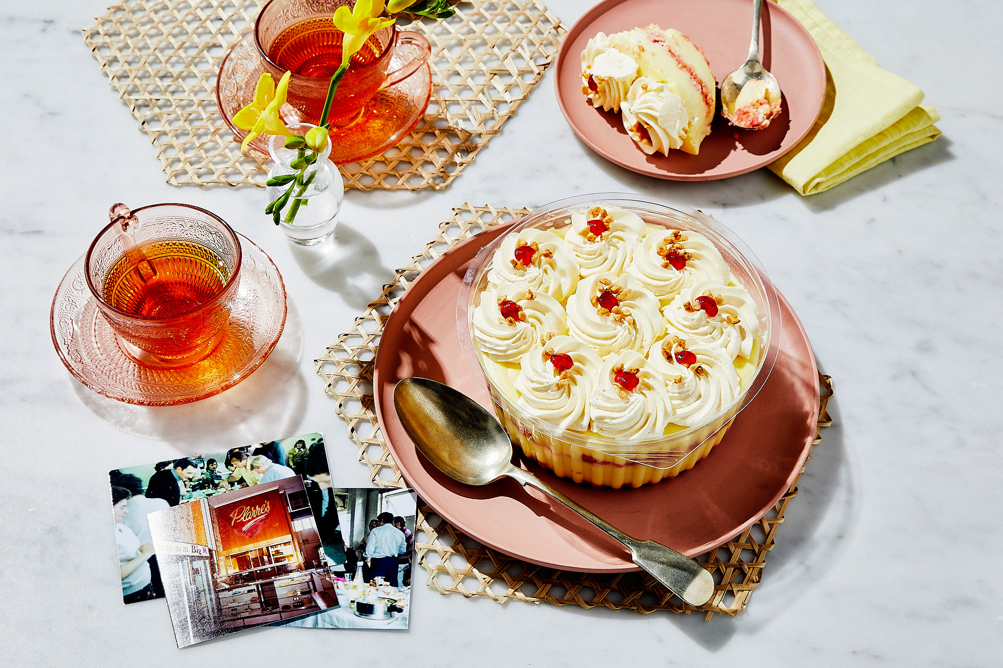 Overhead shot of a cake, teacups with tea, flowers, and photos, all arranged on a marble table, highlighting a food and beverage theme.