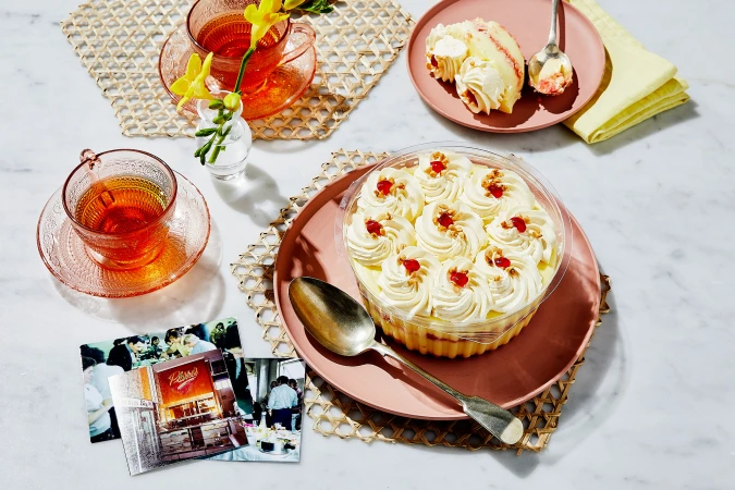 Overhead shot of a cake, teacups with tea, flowers, and photos, all arranged on a marble table, highlighting a food and beverage theme.