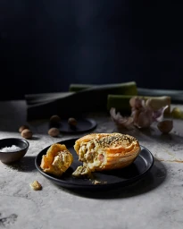 A savory baked pie cut open on a black plate, sitting on a textured stone surface with leeks and garlic in the background.