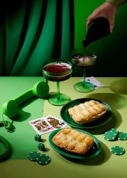 Green still life scene with poured drinks in ribbed glasses, two savory pies on plates, playing cards, dice, and a green telephone.