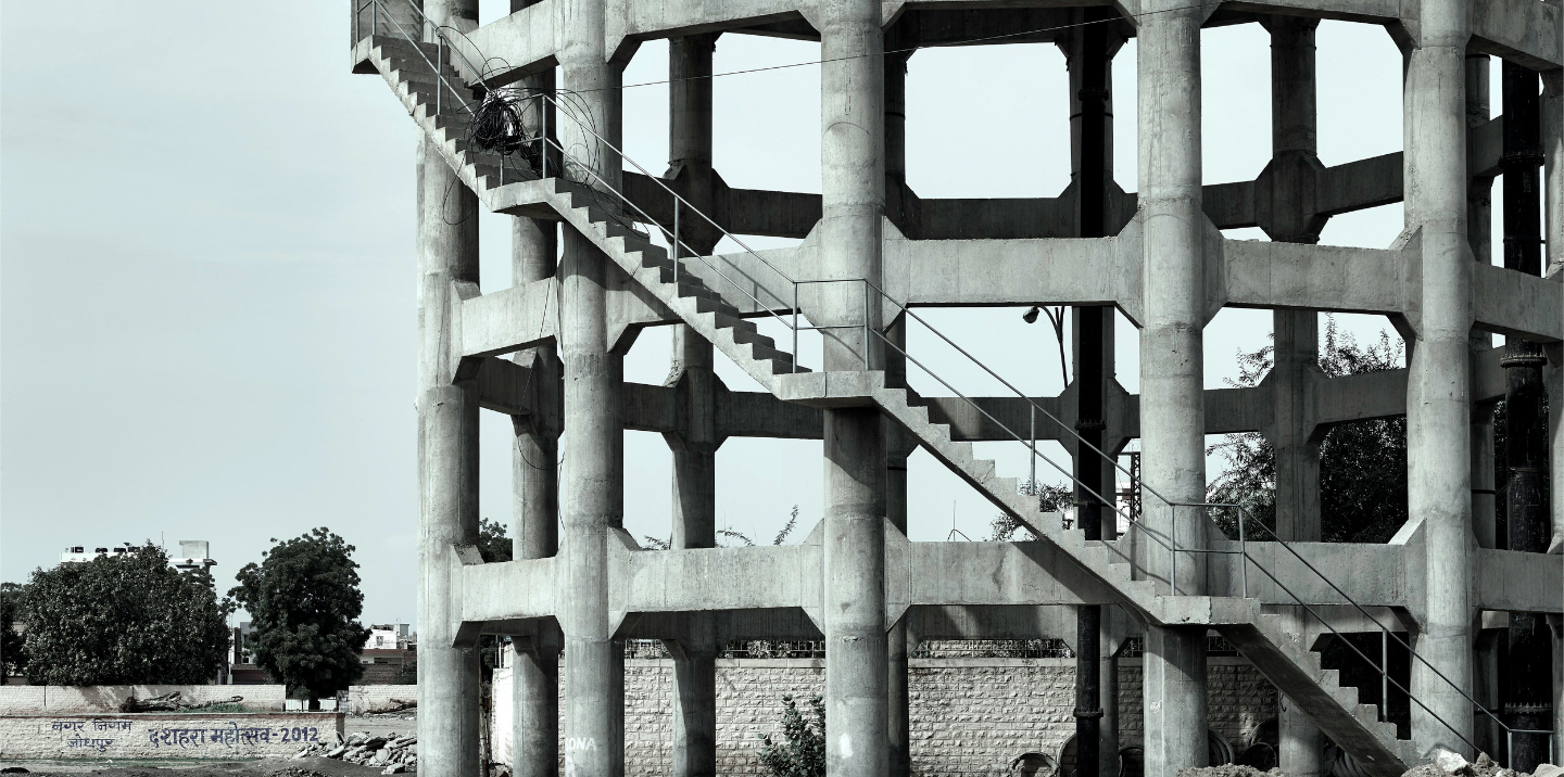 Black and white architectural shot of a tall, unfinished concrete water tower with open framework and staircase rising along one side.