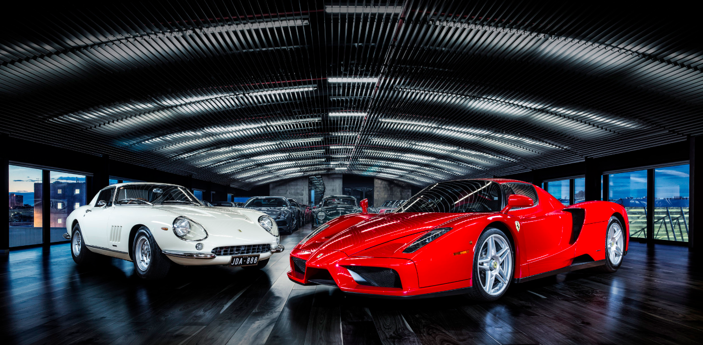 Luxury car collection featuring a red Ferrari and a white classic car in a dark, modern garage under bright overhead lighting.