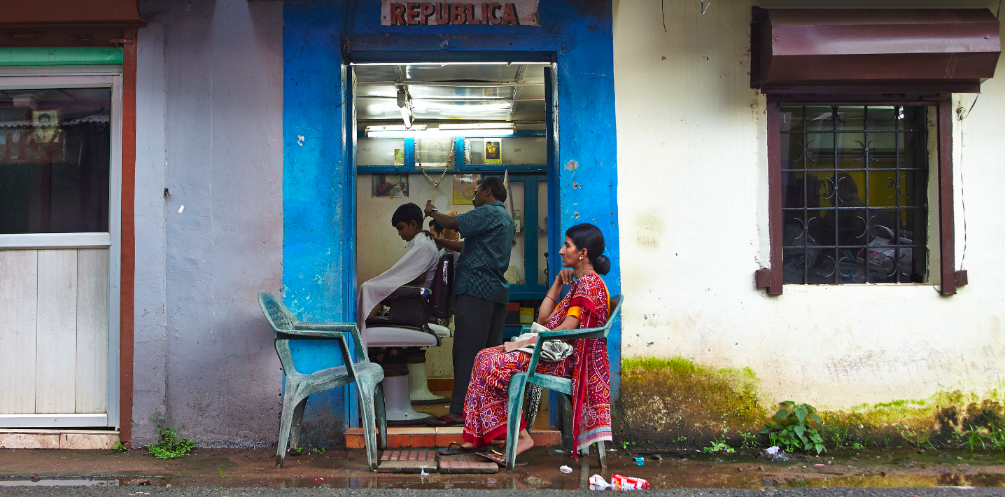 Outdoor scene: barber giving a haircut to a young boy inside a barber shop, with a woman sitting and waiting outside the shop.