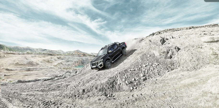 Black pickup truck driving on a rocky landscape against a cloudy sky, with a blue pond visible in the background. 