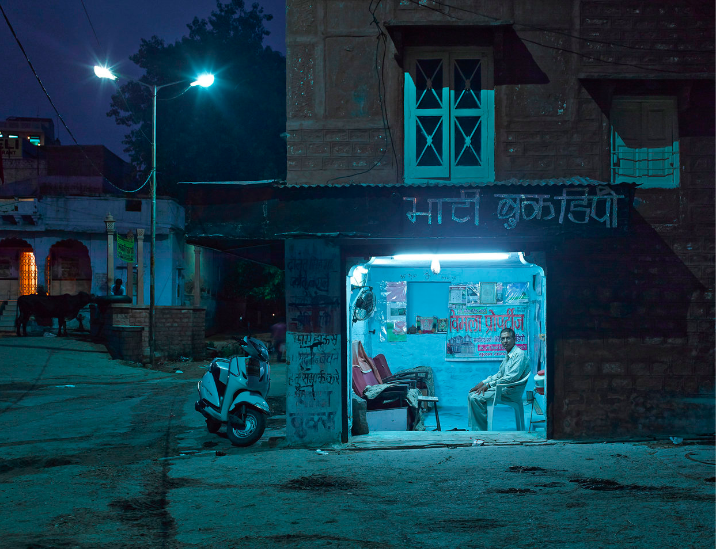 Night scene in India features a man in a shop, scooter parked outside, and buildings lit by streetlights, creating an urban atmosphere.