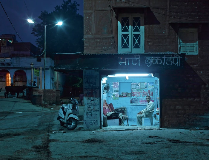 Man seated inside a brightly lit storefront at night on a dark street with a parked scooter.