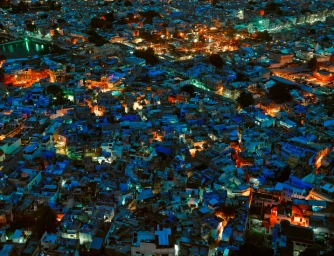 Aerial night view of a dense cityscape illuminated by warm and cool lights across rooftops and narrow streets.