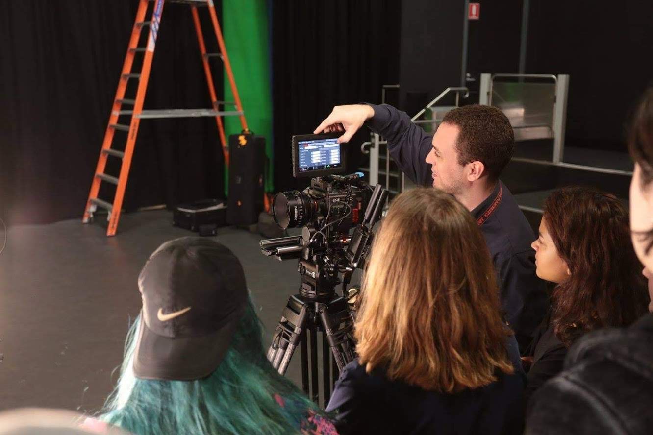 Film crew and students gathered around a camera on a tripod in a studio, with a man adjusting settings on the camera's monitor.