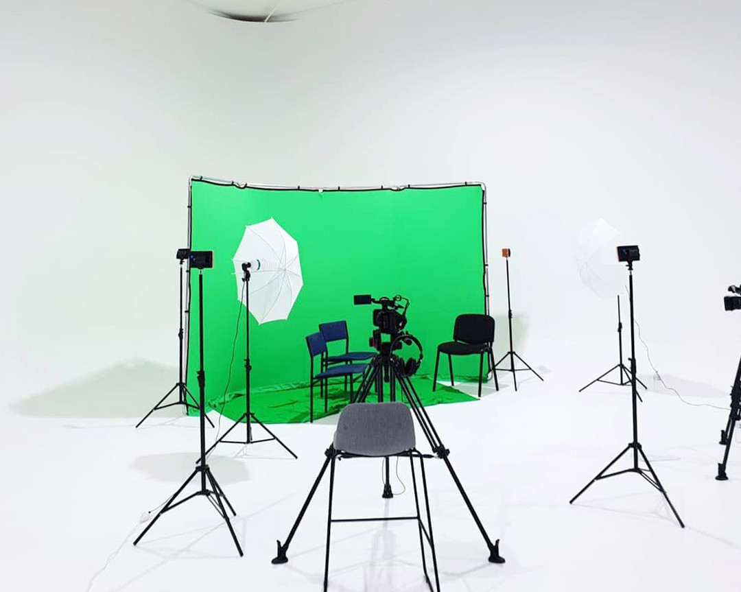 Bright, all-white photo studio features a camera on a tripod, lighting equipment, chairs, and a green screen backdrop for visual production.