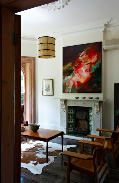 Living room interior with a coffee table on a cowhide rug, fireplace with artwork above mantel, and wooden chairs on a wood floor.