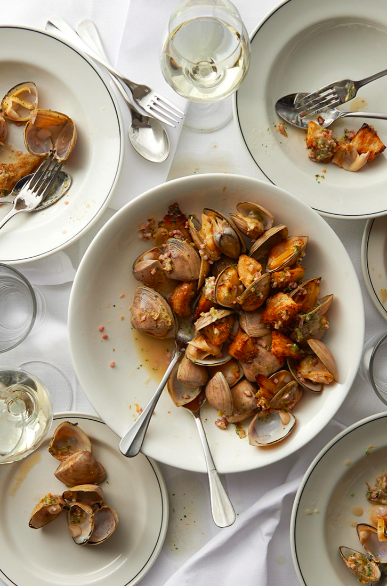 Overhead shot of white plates of clam dishes and a wine glass set on a white tablecloth, evoking a sophisticated dining atmosphere.