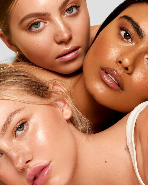 Close-up portrait of three multiethnic young adult women with clean glowing skin, and neutral makeup. Beauty shot with minimal studio background.