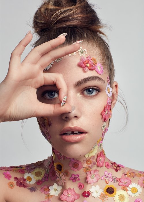 Close-up of a fair-skinned young adult woman adorned with colorful flowers on her face and upper body, making an O.K. sign with her hand.