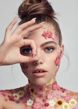Close-up portrait of a woman with pressed flowers adorning her face and neck, framing her eye with hand