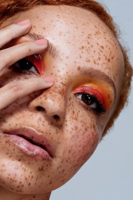 Close-up of a woman's face with freckles, vibrant orange and red eyeshadow, a glossy lip, and her hand partially covering her face.