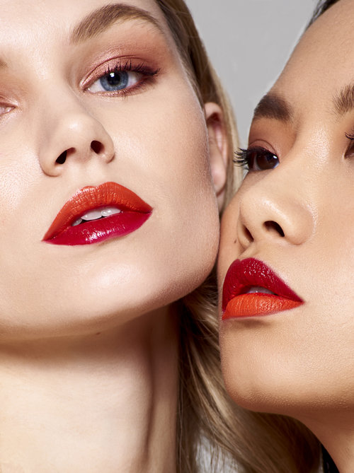 Close-up studio shot of two women with vibrant orange and red ombre lips and complementary makeup, showcasing beauty and fashion.