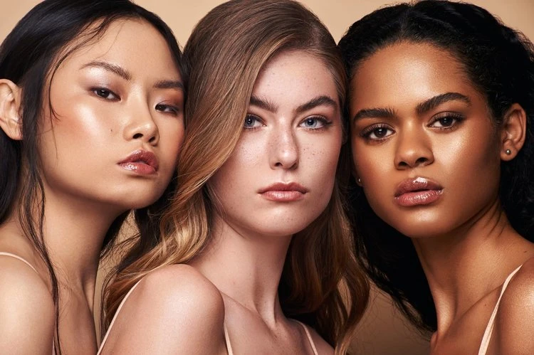 Close-up studio shot of three multi-ethnic young adult women with fair skin, perfect makeup, and long hair, posing against a beige background.