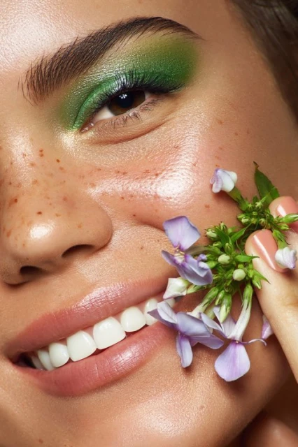 Close-up of a woman's face showcasing green eyeshadow and freckles, with delicate purple flowers accenting her cheek.