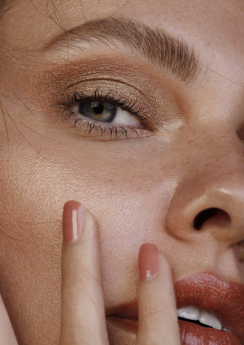 Close-up portrait of an adult female with bronze eyeshadow and coral lipstick, touching her face with manicured nails.