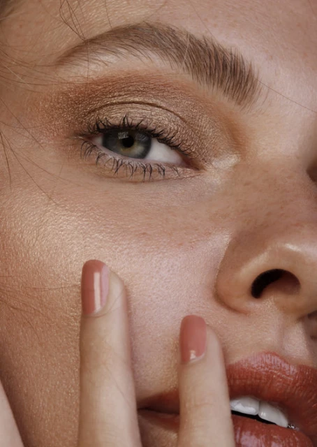 Close-up portrait of an adult female with bronze eyeshadow and coral lipstick, touching her face with manicured nails.