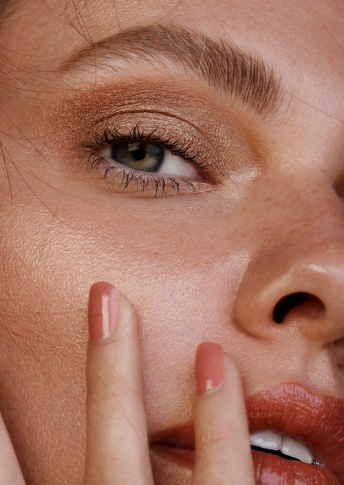 Extreme close-up beauty portrait featuring shimmering copper eyeshadow, freckled skin, and glossy coral-toned lips.