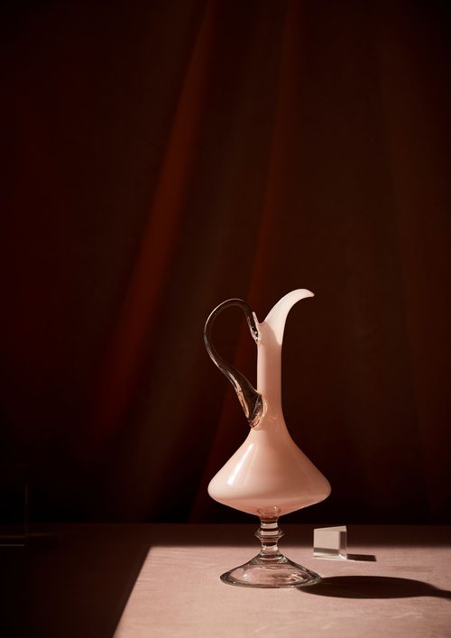 Elegant studio shot of a tall, clear glass and opaque white decanter, set on a light surface with soft brown background and shadows.