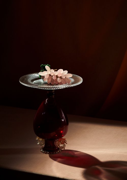 Still life photograph of a bunch of pale pink grapes on a clear glass plate and deep red glass pedestal with dark brown background.