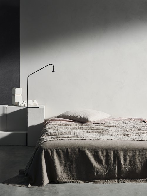 Minimalist bedroom interior featuring a bed with a beige blanket, a bedside table with stacked blocks, and a modern lamp against a white wall.
