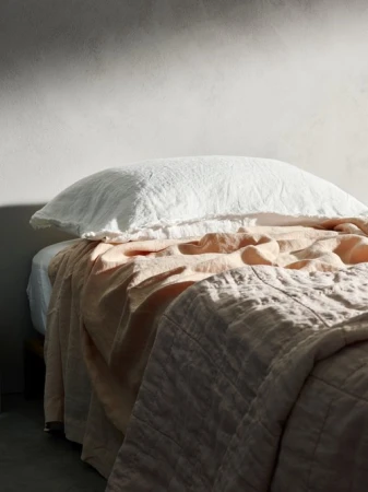 Bedroom interior with a bed featuring a white pillow, pink sheets, and a textured grey comforter. Natural light softly illuminates the setting.