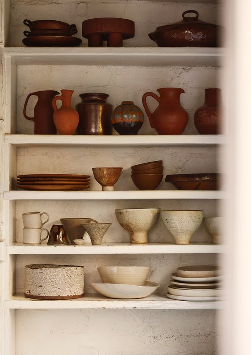 Still life photo of rustic pottery and dishes arranged on shelves, showcasing neutral tones and textures in a homey kitchen setting.