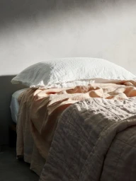 Close-up of a made bed featuring a white linen pillow and peach-toned bedding against a neutral textured wall.