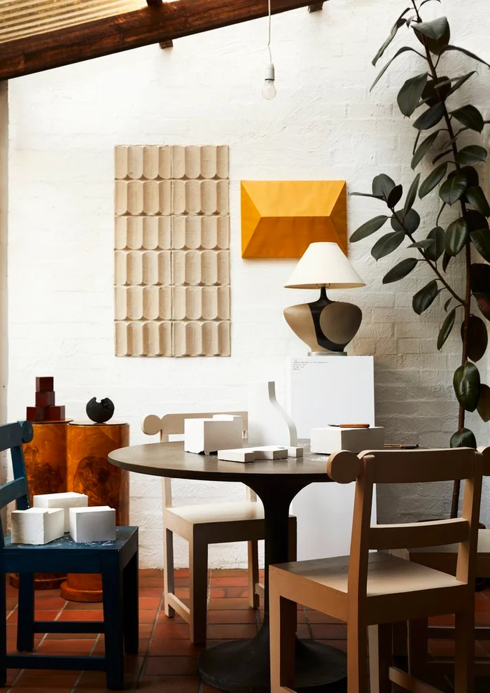 Interior styling featuring a round dark wooden table, multiple chairs, a sculptural lamp, and decorative art objects on white brick walls.