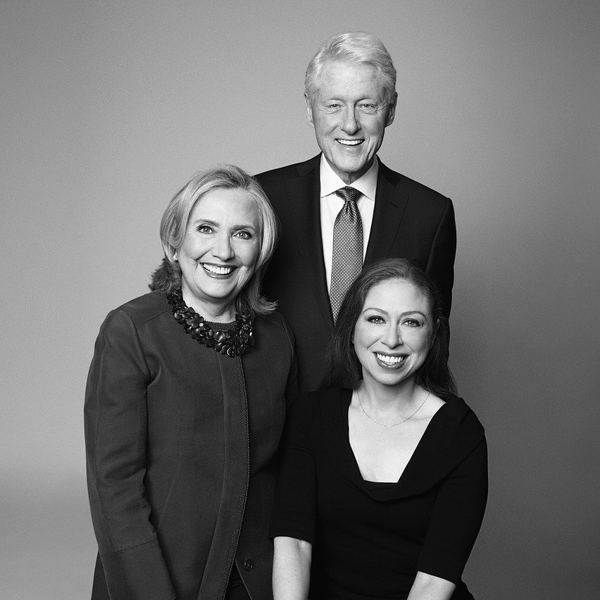 Three people smiling in a black and white studio portrait: Hillary Clinton, Bill Clinton, and Chelsea Clinton are posing indoors.