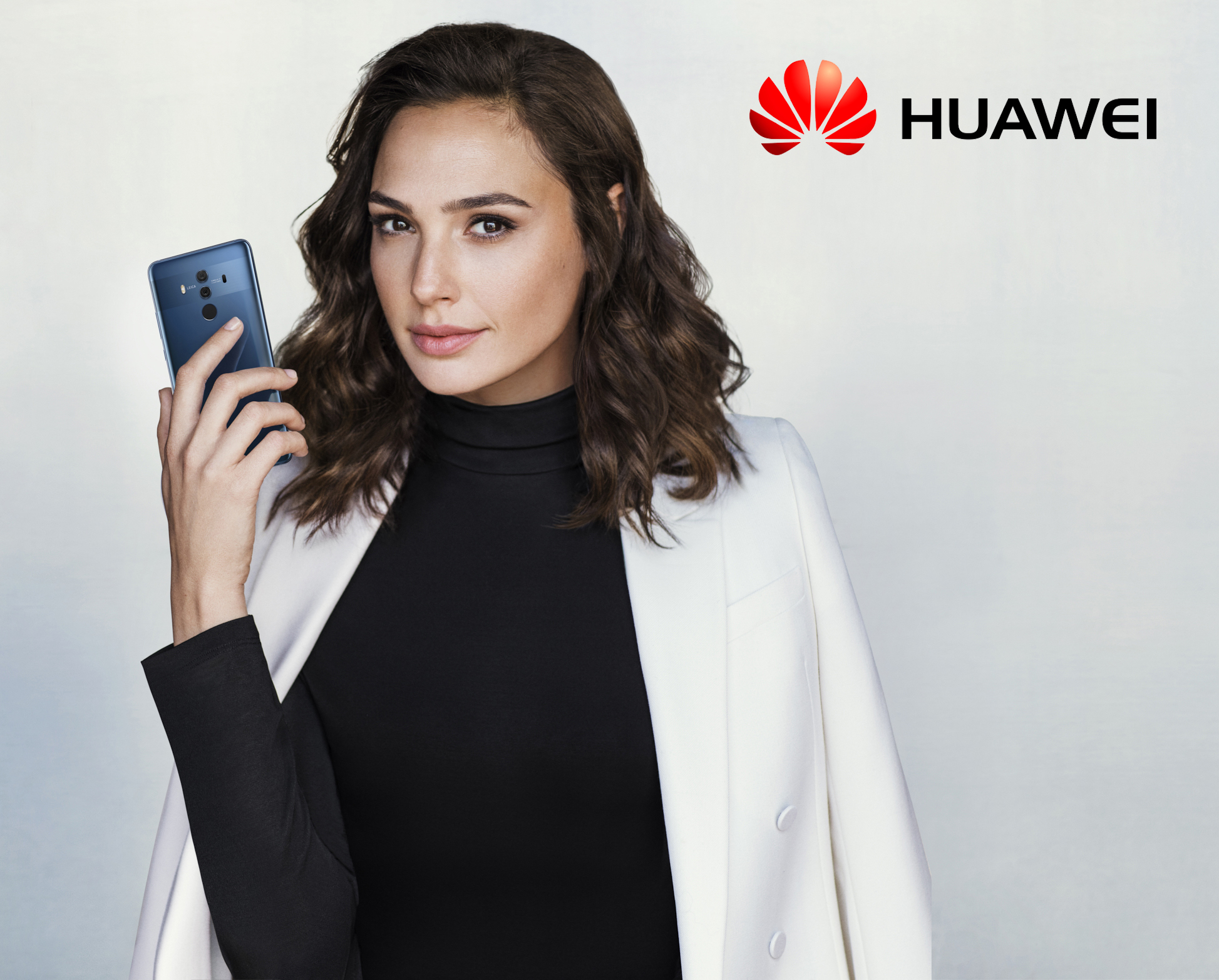 Gal Gadot holds a blue smartphone against a white background, wearing a black turtleneck and white blazer, with Huawei logo visible.