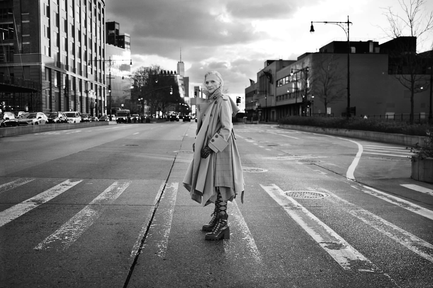 Monochrome portrait of an adult female in a stylish coat and boots crossing an urban street in a city setting.