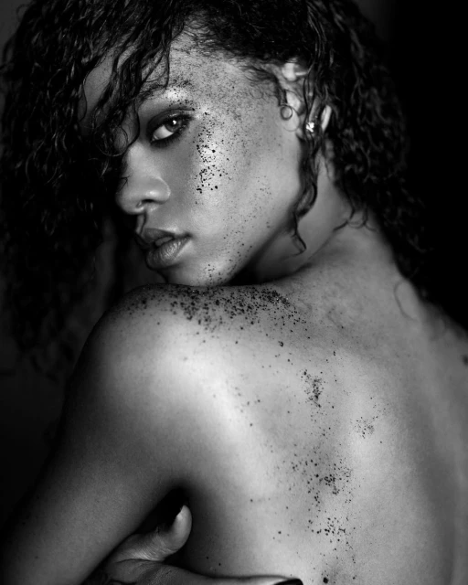 Black and white portrait of a young adult female with dark curly hair, looking over her shoulder with dark specks on her face and back.