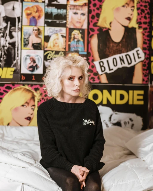Blondie singer Debbie Harry in black sweater sitting in bed in front of wall covered in Blondie posters and album covers.