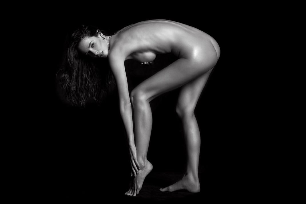 Monochrome portrait of an adult woman bending forward, lit with soft light against a dark background, revealing her pose and physique.