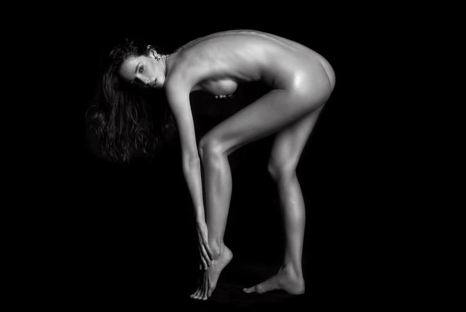Monochrome portrait of an adult woman bending forward, lit with soft light against a dark background, revealing her pose and physique.