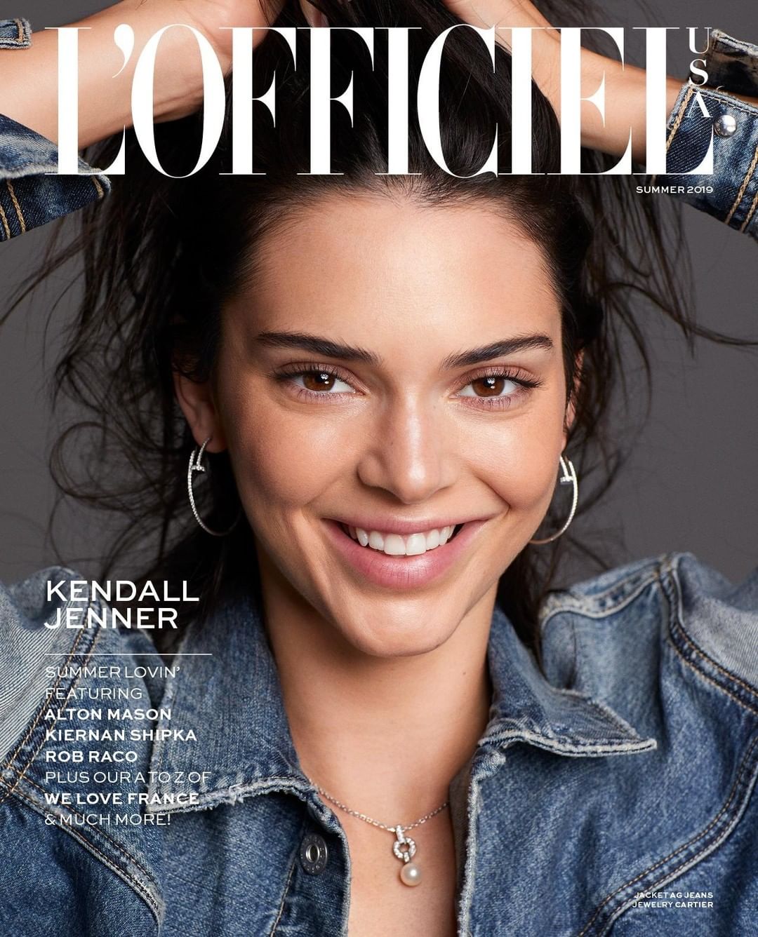 Close-up magazine cover shot of smiling Kendall Jenner wearing a denim jacket and silver hoop earrings against a grey background.
