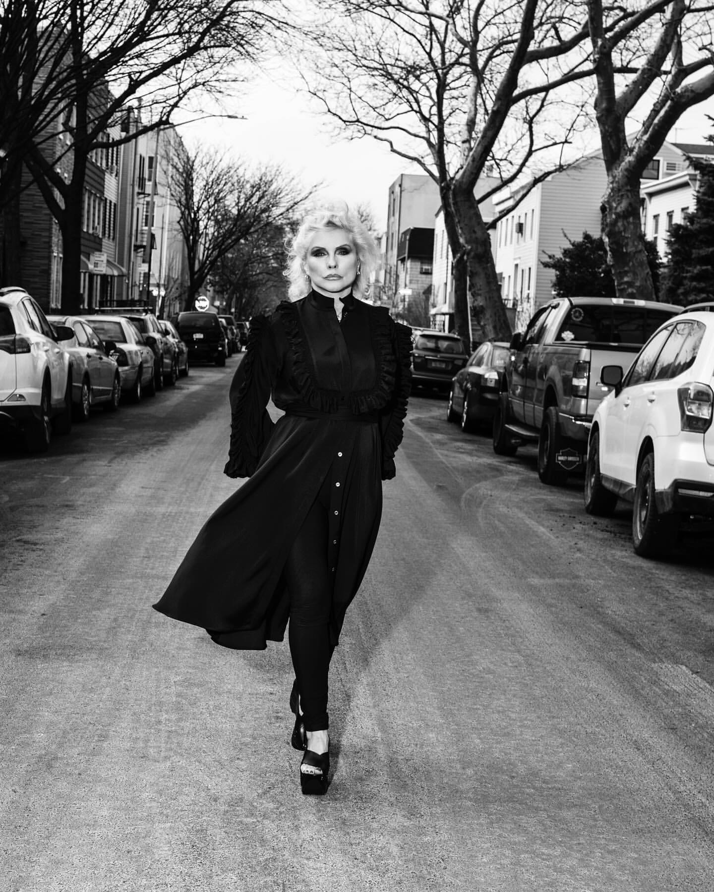 Debbie Harry poses in a black dress with ruffles on a street lined with parked cars and trees in a black-and-white outdoor portrait.