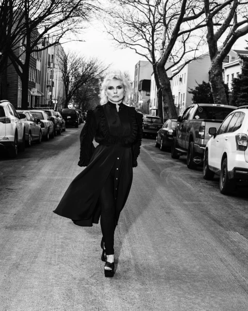 Debbie Harry poses in a black dress with ruffles on a street lined with parked cars and trees in a black-and-white outdoor portrait.