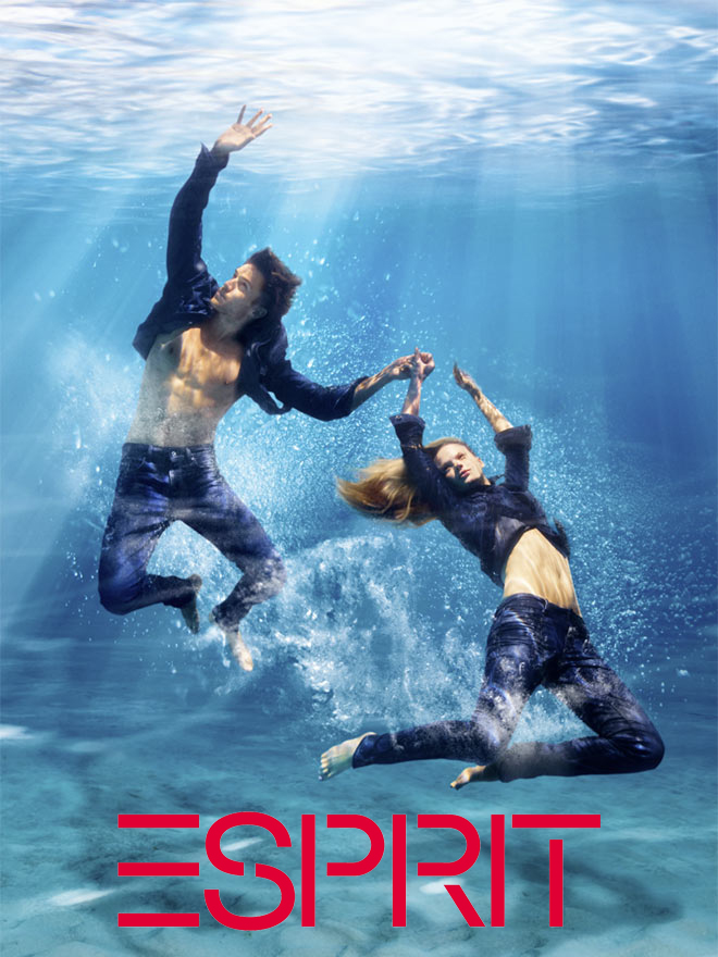 A man and a woman wearing jeans and jackets are underwater with light shining through the clear water above them.