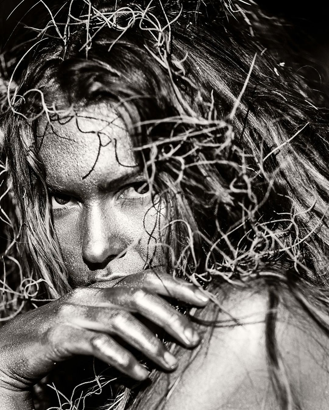 Monochrome close-up of an adult woman with long hair and silver body paint, partially concealing her face with a silver-painted hand.